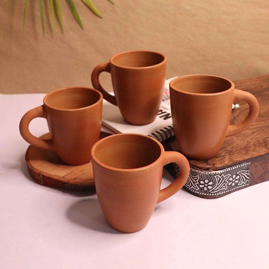A collection of four handcrafted red terracotta mugs by Vins Art House arranged on a white and wooden surface.